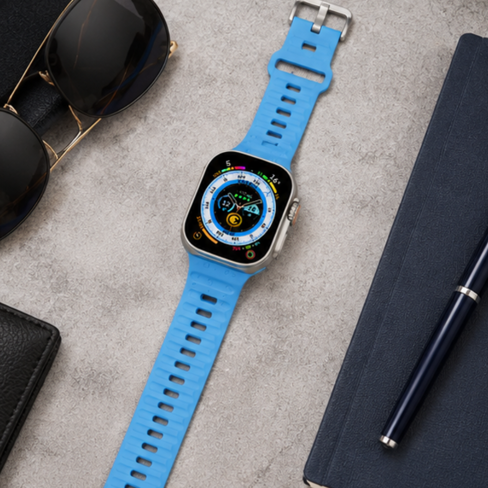 Apple Watch Ultra with a bright blue rubber sport band placed in a flat lay with sunglasses, a notebook, and a pen on a gray background