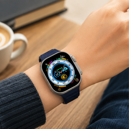 Person wearing an Apple Watch Ultra with a navy blue sport rubber band on their wrist, shown in a casual lifestyle setting with a wooden table in the background