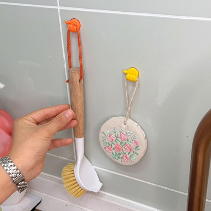 Hand holding a kitchen brush with a floral decoration on a tiled wall.